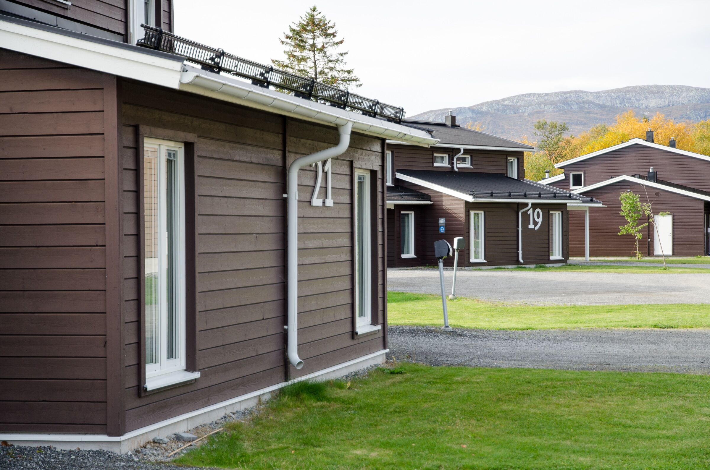 A row of brown wooden houses with white trim, numbered 19, in a residential area with a mountain backdrop.