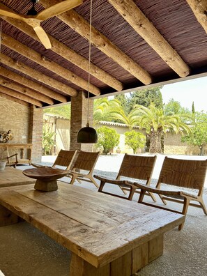 Un coin repas en bois, avec une grande table et quatre chaises en osier, un luminaire suspendu et une vue sur un jardin agrémenté de palmiers et d’un mur en pierre.