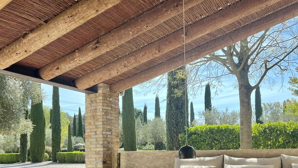 An outdoor seating area with wooden tables and chairs, a cushioned sofa, and a view of a garden with trees and a clear sky. - Spectacular unique villa in the Emporda (Calabuig)