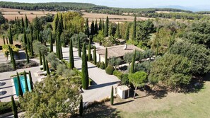 Vue aérienne d’une propriété dont le bâtiment principal est entouré d’un jardin bien entretenu, agrémenté de grands cyprès, d’une piscine et d’un petit abri de jardin.