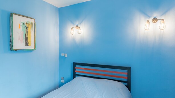 Ein Zimmer mit blauen Wänden, einem Bett mit weißer Matratze und einem gestreiften Kopfteil, sowie zwei Wandleuchten.