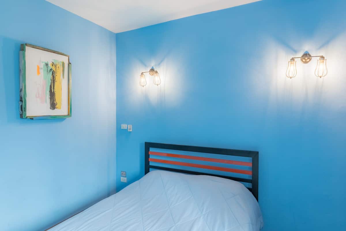 Une pièce aux murs bleus, avec un lit doté d’un matelas blanc et d’une tête de lit à rayures, ainsi que deux appliques murales.