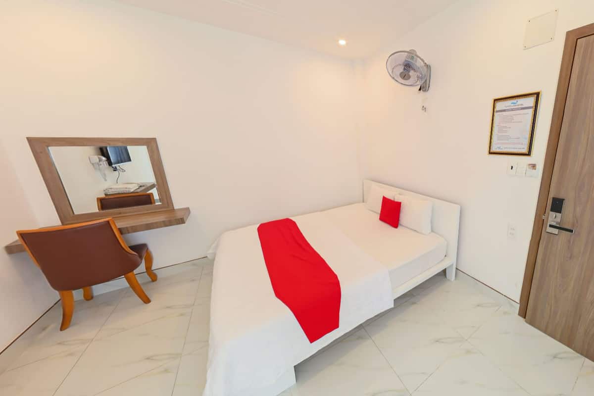 A bedroom with a bed, a desk, a chair, a mirror, a wall-mounted fan, and a door.