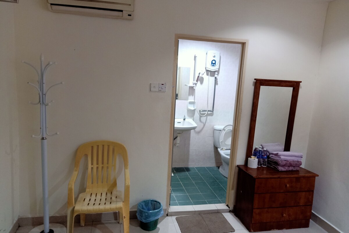 A bathroom with a toilet, sink, and shower. There is a wooden chair, a blue trash bin, and a wooden dresser with a mirror.