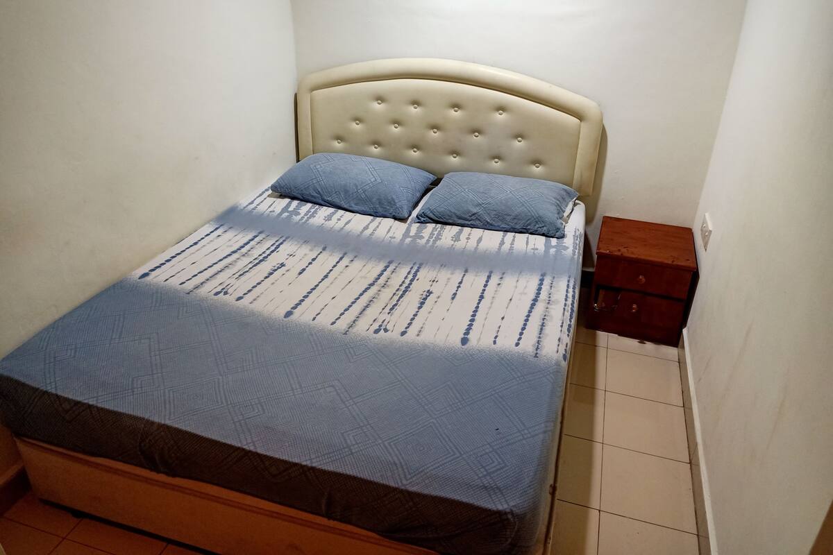 A small, neatly made bed with a tufted headboard and two pillows, a wooden nightstand with a drawer, and a tiled floor.