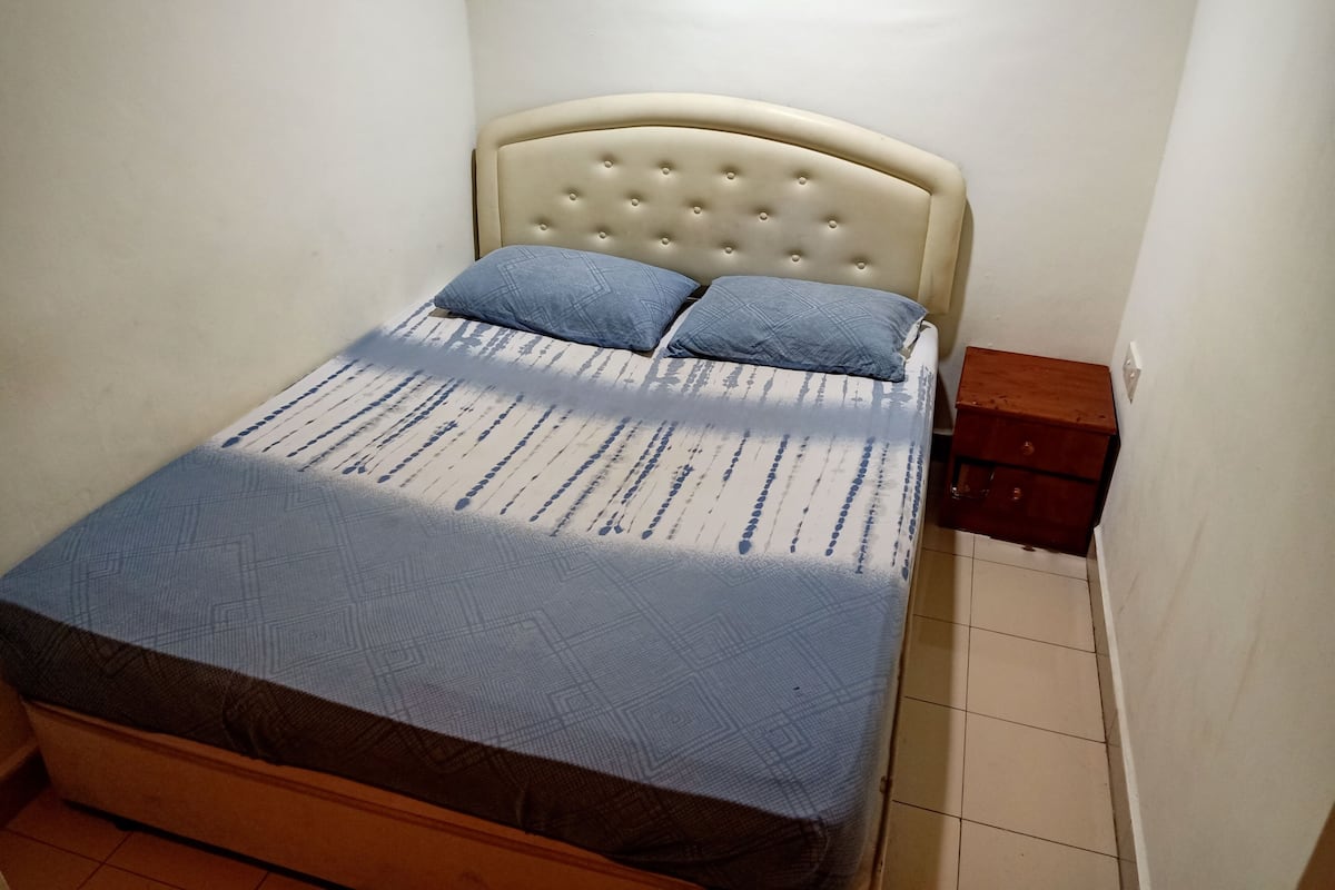 A small, neatly made bed with a tufted headboard and two pillows, a wooden nightstand with a drawer, and a tiled floor.