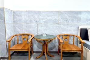 Habitación de esquina con dos sillas de madera y una mesa de vidrio, televisor y pared de azulejos.