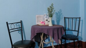 Una habitación pequeña con pared azul y blanca, piso de baldosas azules, una mesa con mantel morado estampado con lunares, dos sillas negras y un jarrón blanco con una planta.