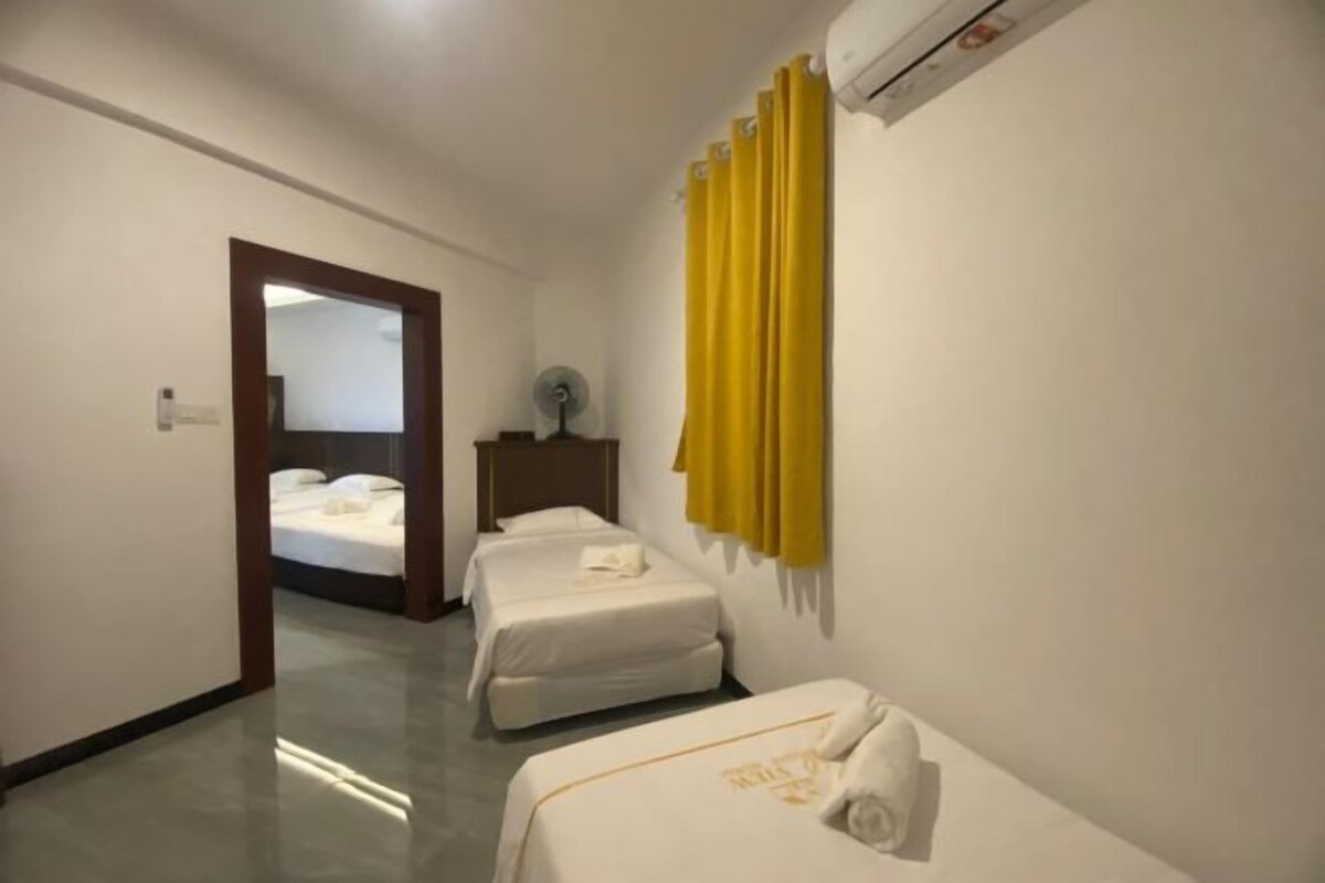 Quarto de hotel com duas camas, uma mesa de cabeceira, uma luminária e uma janela com cortinas.