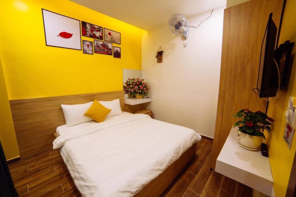 A hotel room with a wooden headboard, a bed with white and yellow bedding, a bedside table with a vase of flowers, and a wall-mounted fan.