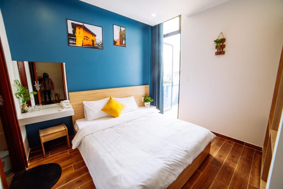 A hotel room with a large bed, a wooden headboard, a bedside table with a mirror, a small plant, and a window with curtains.