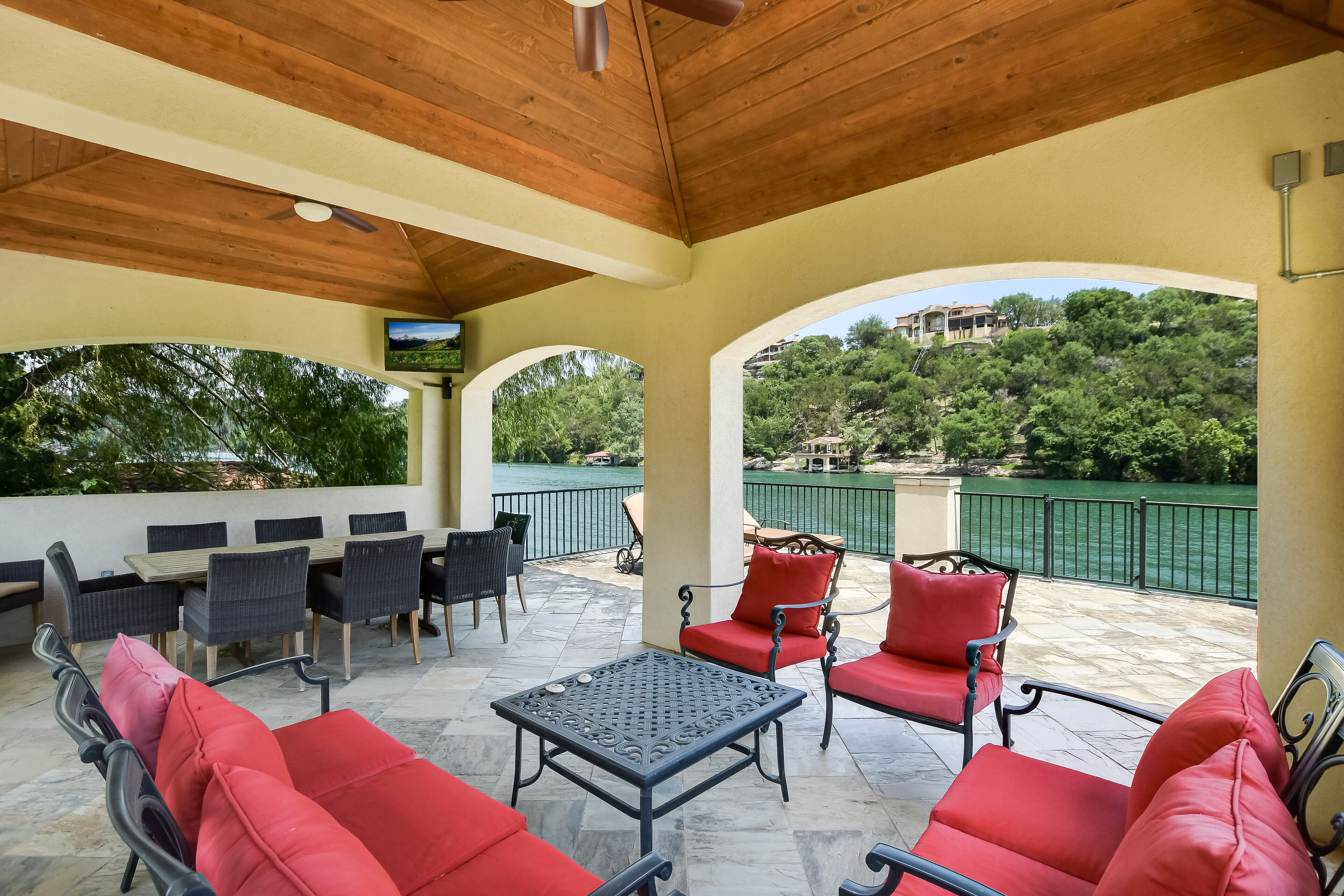 A covered patio with red and black furniture, a TV mounted on the wall, and a view of a green landscape and water.