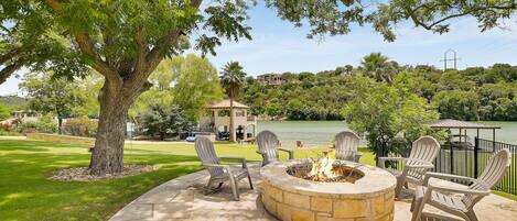 A patio with a stone fire pit and Adirondack chairs, overlooking a lake and a house in the background.
