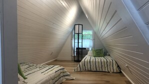 Une chambre mansardée chaleureuse, avec un plafond en pente, un lit recouvert d’une couette à rayures, une petite table ronde et une fenêtre donnant sur un paysage verdoyant.