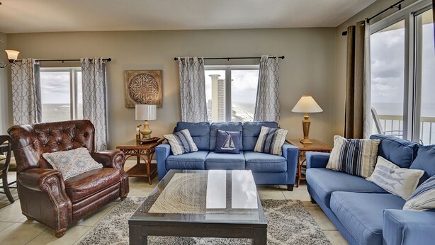 A living room with a brown leather armchair, a blue sofa set, a glass coffee table, and a view of the city through large windows. - Calypso Resort by BBG (Panama City Beach)