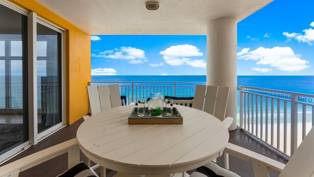 Een balkon met een tafel gedekt voor twee personen, met uitzicht op een strand met een helderblauwe lucht en zee.