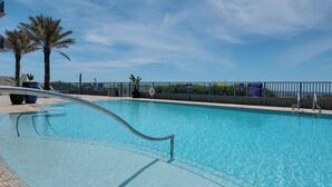En pool med klart vatten och ett metallräcke runt om, omgiven av ett staket med skyltar, palmer och en klar himmel.
