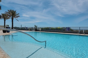 Une piscine aux eaux cristallines, dotée d’une rambarde métallique, entourée d’une clôture, de panneaux indicateurs, de palmiers et sous un ciel dégagé.