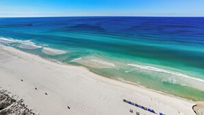 Ein Strand mit klarem türkisblauem Wasser, einer Mole, die ins Meer hinausführt, und einer Reihe blauer Liegestühle entlang der Küste.