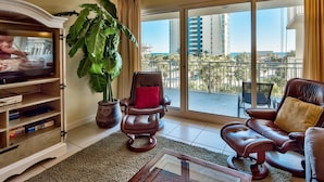 A living room with a television, a large plant, a leather sofa, and a view of a cityscape through large windows. - Sterling Shores 303 (Destin)