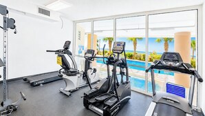 Ein Fitnessraum mit Laufbändern, Ellipsentrainern und Steigern, mit Blick auf ein Schwimmbecken und eine Palmenlandschaft sowie den Ozean.