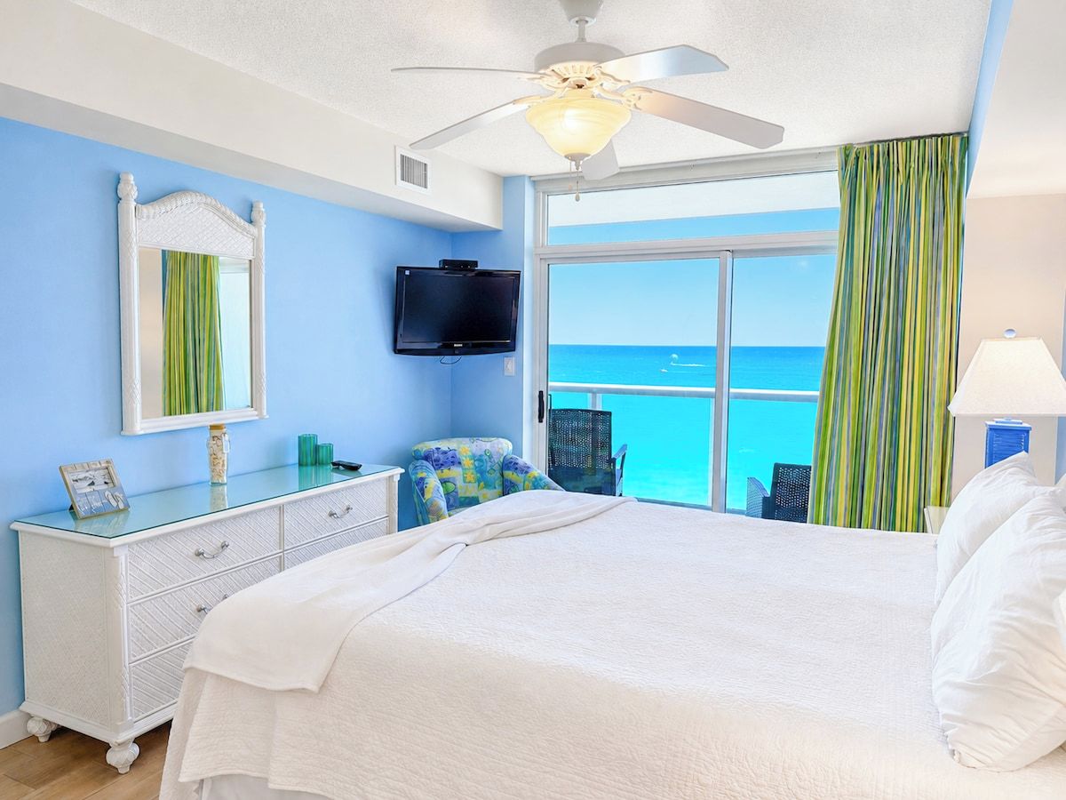 A hotel room with a bed, a ceiling fan, a TV, a window with a view of the ocean, and a dresser with a mirror.