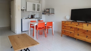 A compact living space with a kitchenette, a dining area with orange chairs, a wooden table, a TV on a wooden cabinet, and a geometric ceiling light.