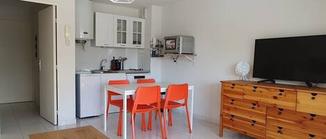 A compact living space with a kitchenette, a dining area with orange chairs, a wooden table, a TV on a wooden cabinet, and a geometric ceiling light.