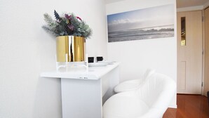 Un baño blanco con una silla blanca, un jarrón dorado con flores y un cuadro de una playa en la pared.