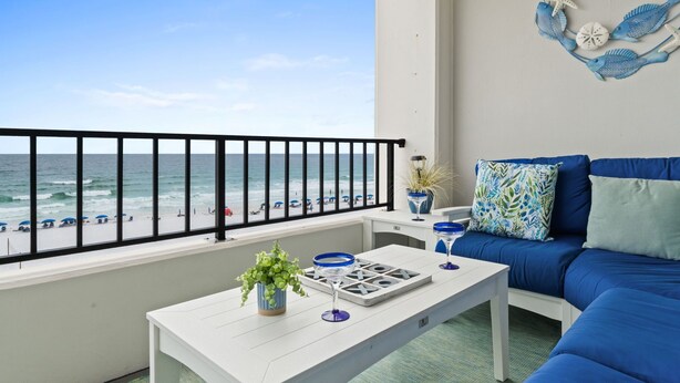 A balcony with a view of the beach, a white table with a plant, a blue sofa, and a decorative wall piece. - SD 508: Beachfront Boho - Indulge in Sunset Serenity with Gulf Views! (Fort Walton Beach)