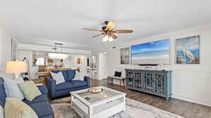 A living room with a blue sofa, a glass coffee table, a ceiling fan, a television, and artwork on the walls. - SD 508: Beachfront Boho - Indulge in Sunset Serenity with Gulf Views! (Fort Walton Beach)