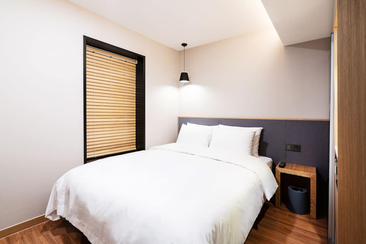 A bedroom with a bed, a bedside table, a lamp, and a window with blinds.
