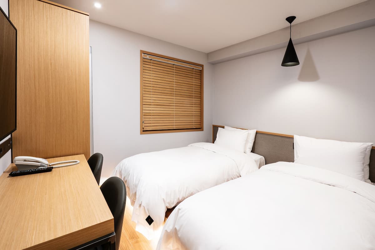 A hotel room with two beds, a desk with a telephone, and a window with blinds.