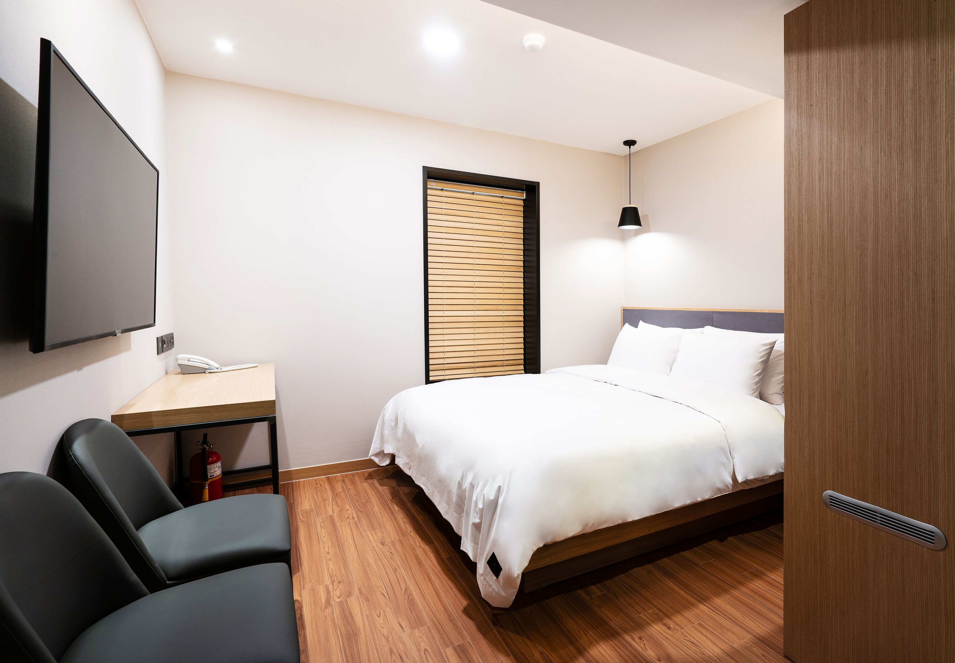 A modern hotel room with a bed, a desk, a chair, a television, and a window with blinds.