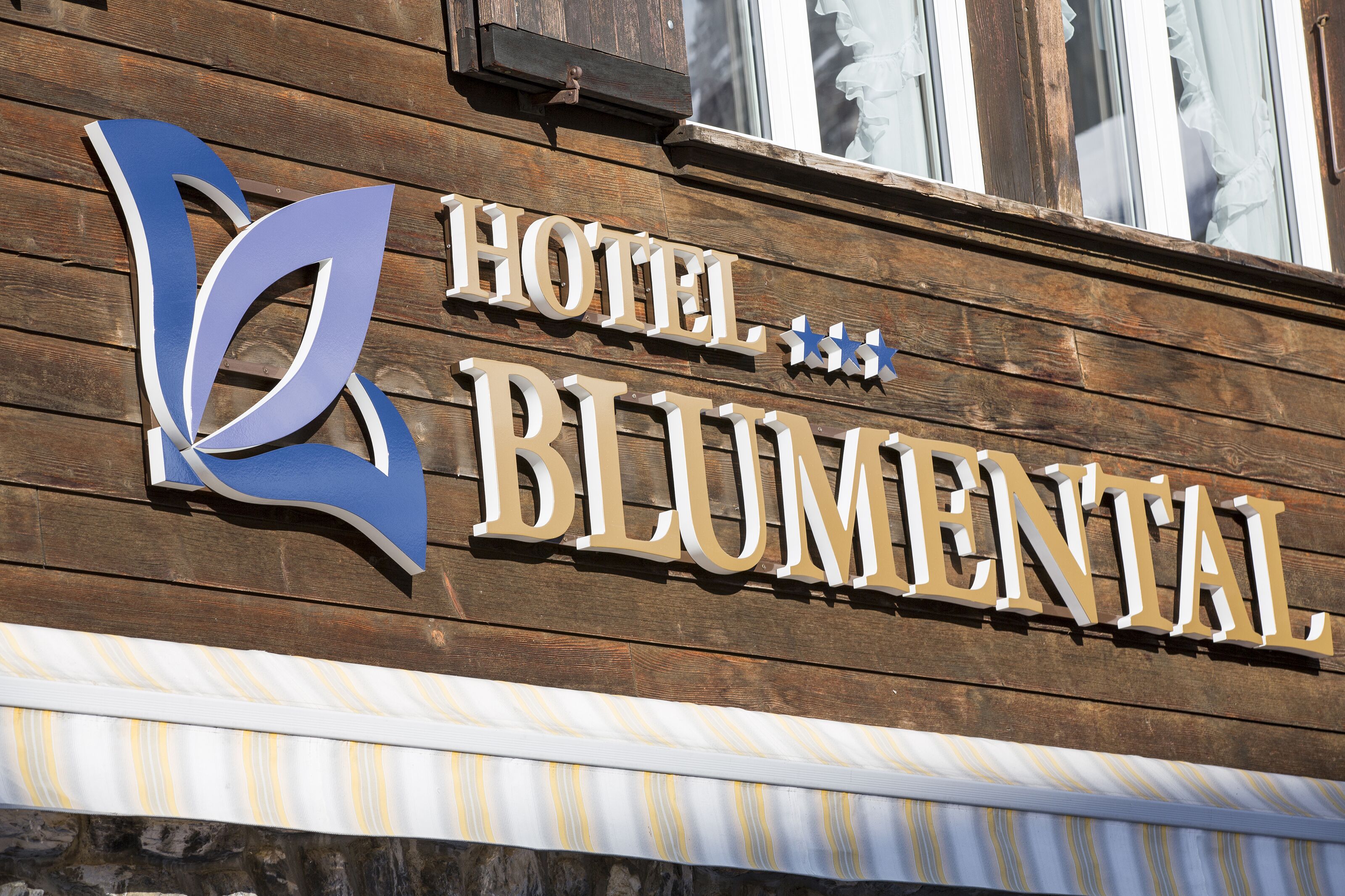 Fachada de madera con el letrero "HOTEL BLUMENTAL" y un logotipo azul.