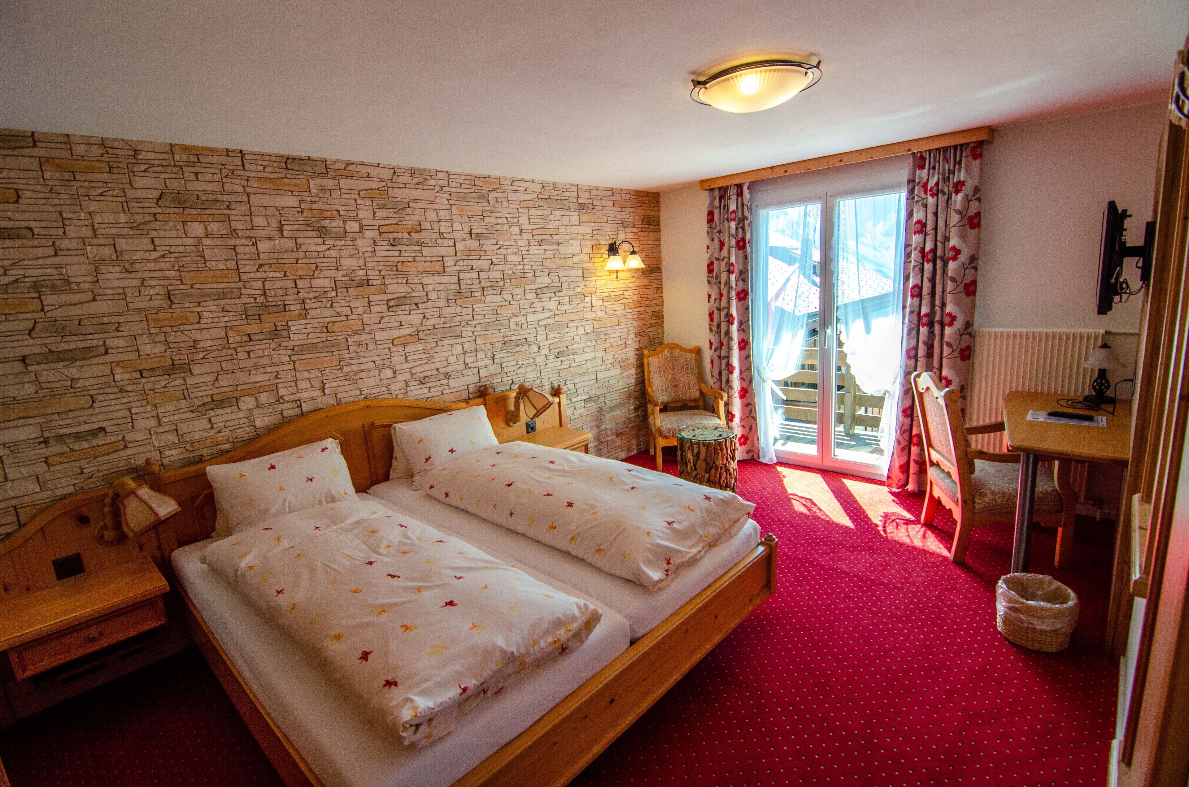 A room with a stone wall, a double bed, a wooden nightstand, a chair, a small table, and a window with curtains.