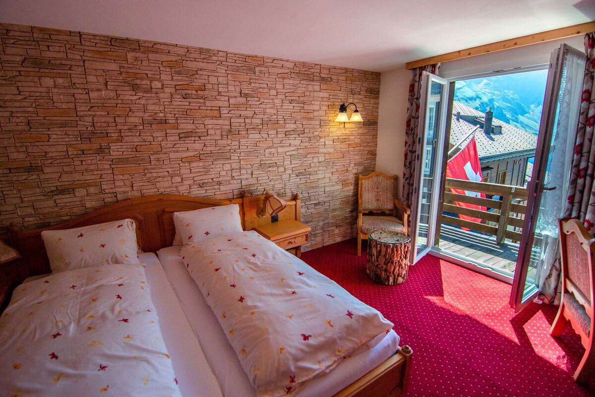A hotel room with a stone wall, a bed with floral bedding, a wooden nightstand, a chair, and a balcony with a view.