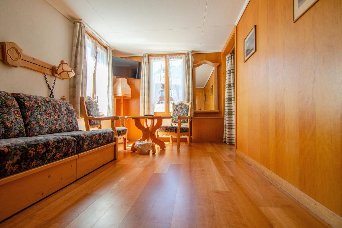 A cozy room with a wooden floor, a floral sofa, a wooden dining table with chairs, and a TV mounted on the wall.