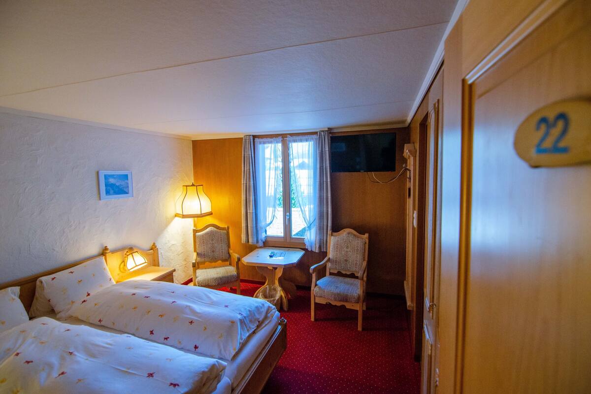 A hotel room with two beds, a small table, two chairs, a TV, and a window with curtains.