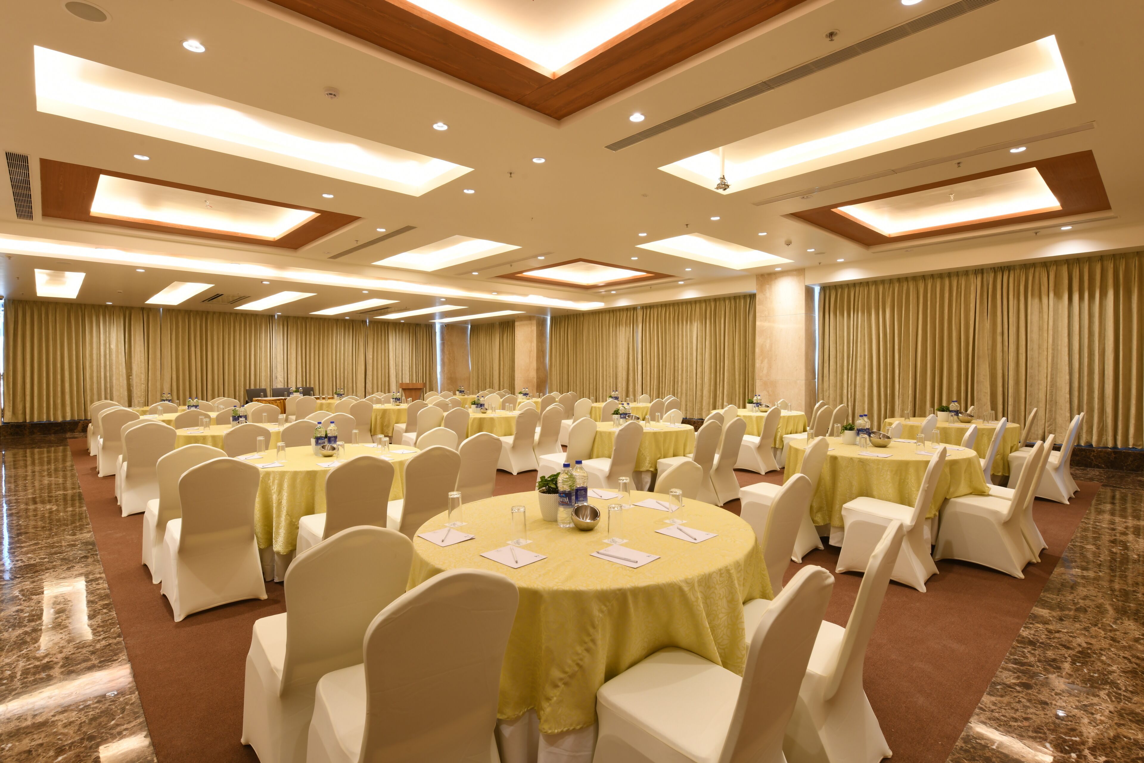 A large conference room with round tables, white chairs, and yellow tablecloths.