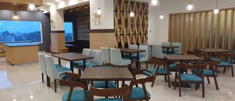A modern dining area with wooden tables and blue cushioned chairs, overhead pendant lighting, and a large screen displaying a cityscape.