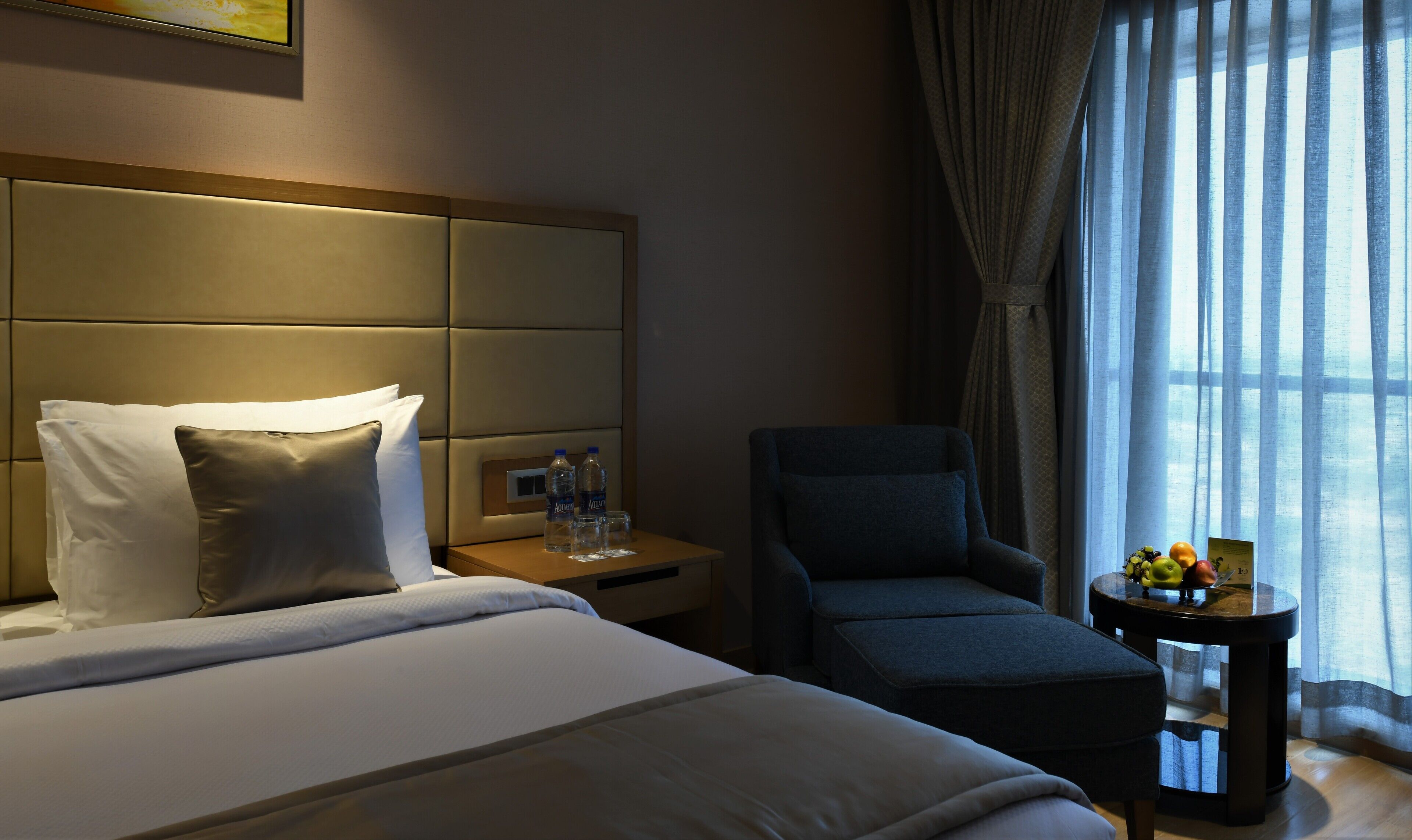 A hotel room with a large bed, a chair, a small table with a fruit bowl, and a window with curtains.