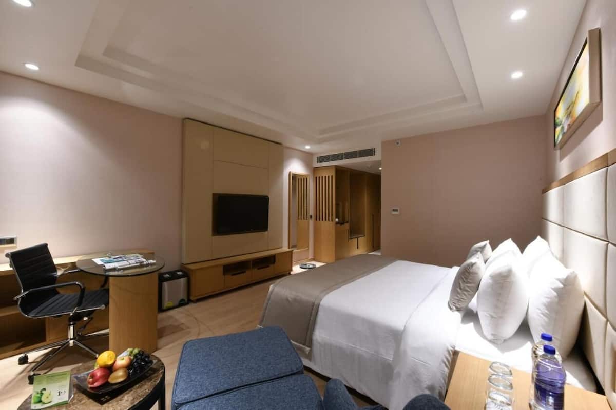 A hotel room with a bed, a desk with a chair, a television, and a small table with fruits and a water bottle.