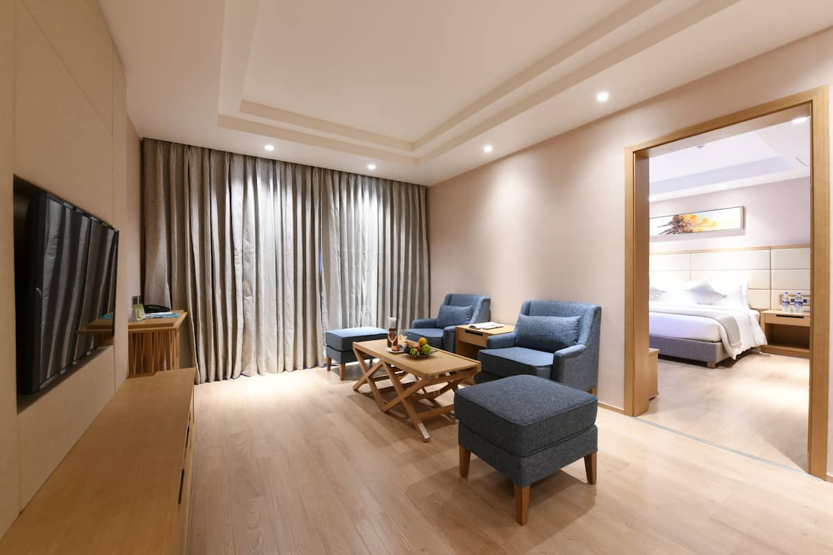 A modern hotel room with a wooden floor, a desk with a chair, two armchairs, a small table with fruit, and a bed with a white headboard.
