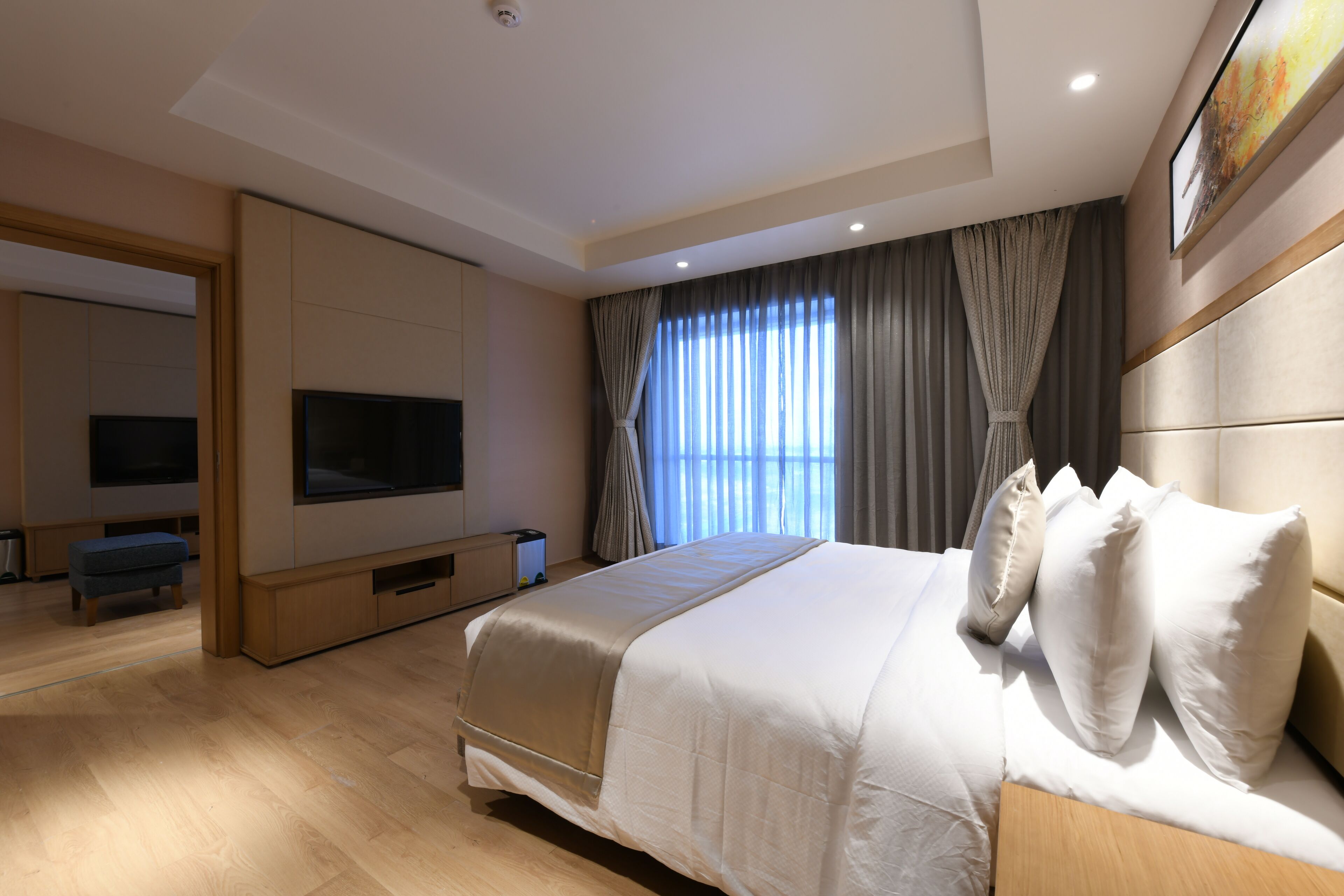 A modern hotel room with a large bed, a flat-screen TV, a wooden bench, and a large window with curtains.