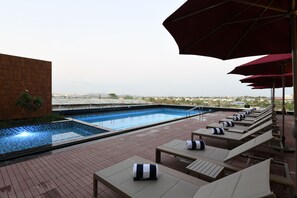 Un espace piscine aménagé sur le toit, avec des chaises longues et des parasols, offrant une vue sur une étendue d’eau et des bâtiments situés au loin.