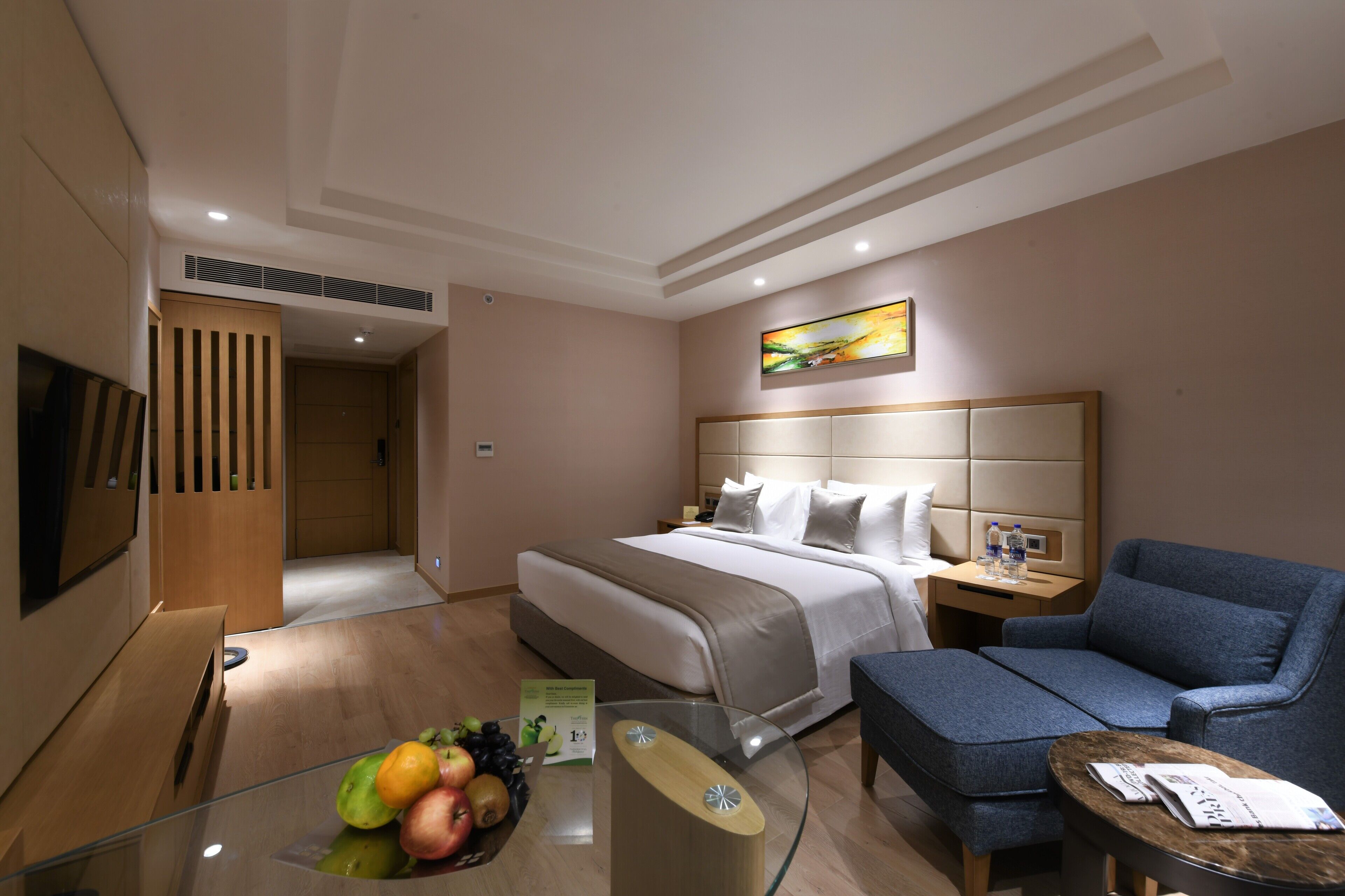 A hotel room with a bed, a sofa, a glass table with fruits, a TV, and a painting on the wall.