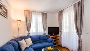 Una sala de estar con un sofá azul, una mesa de centro de vidrio con fruta, y un televisor sobre un mueble de madera.