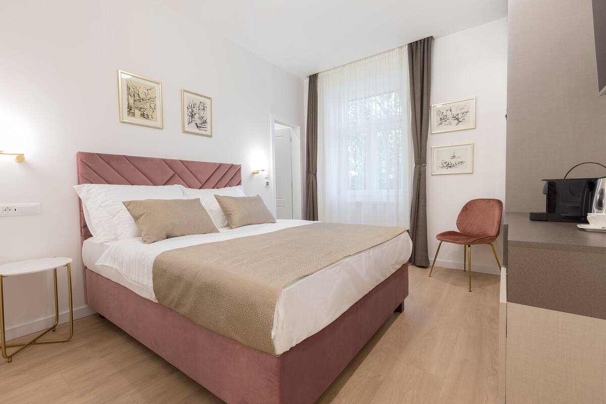 A modern hotel room with a large bed, a small side table, a chair, and a desk with a coffee maker.