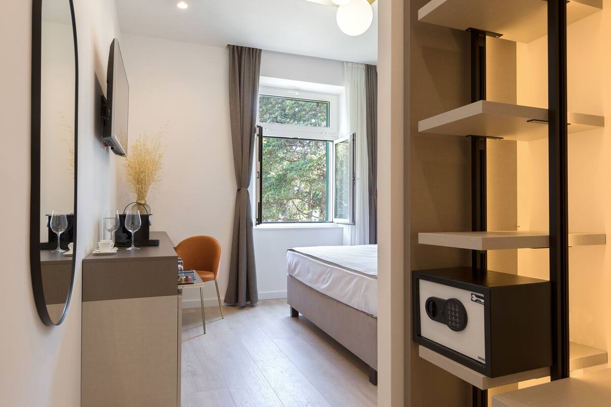 A modern hotel room with a bed, a desk, a chair, a mirror, a window with curtains, and a built-in safe.